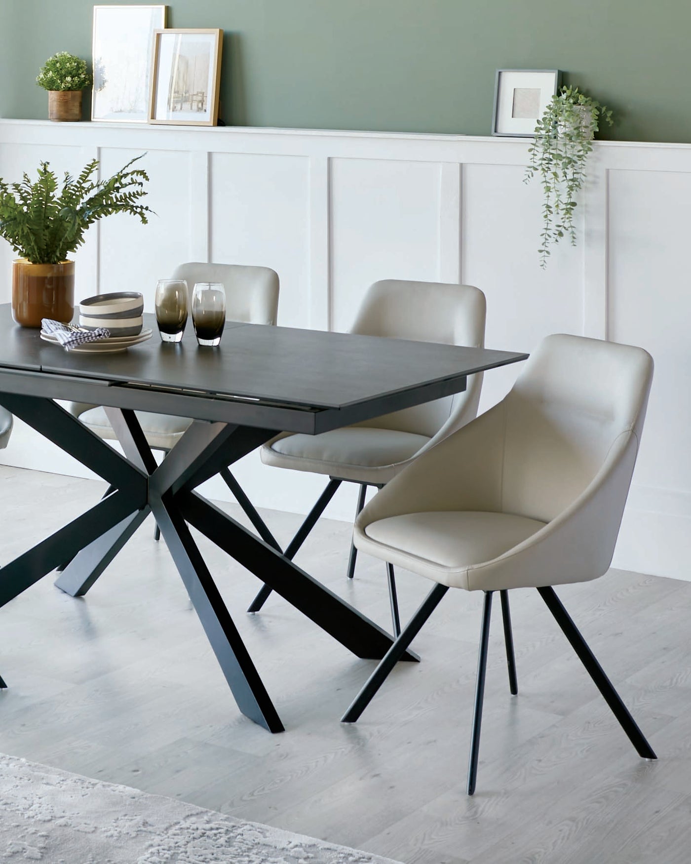 A modern dining set featuring a rectangular table with a dark wood finish and an angular black metal base, paired with three sleek, light grey upholstered chairs with black metal legs. The table is accessorized with a small pot plant, striped bowls, and two dark glasses.