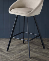 A stylish, tan velvet bar stool with sleek black metal legs against a dark blue panelled wall and wooden floor.