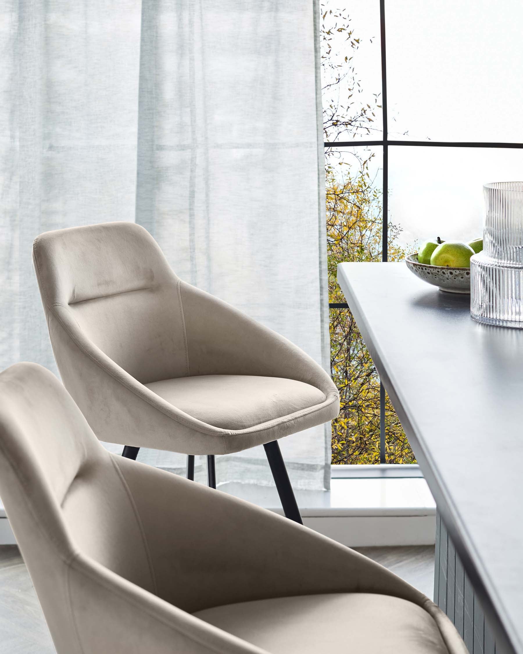 Beige upholstered chairs sit beside a table with fruit and glassware. A large window and sheer curtain provide natural light.
