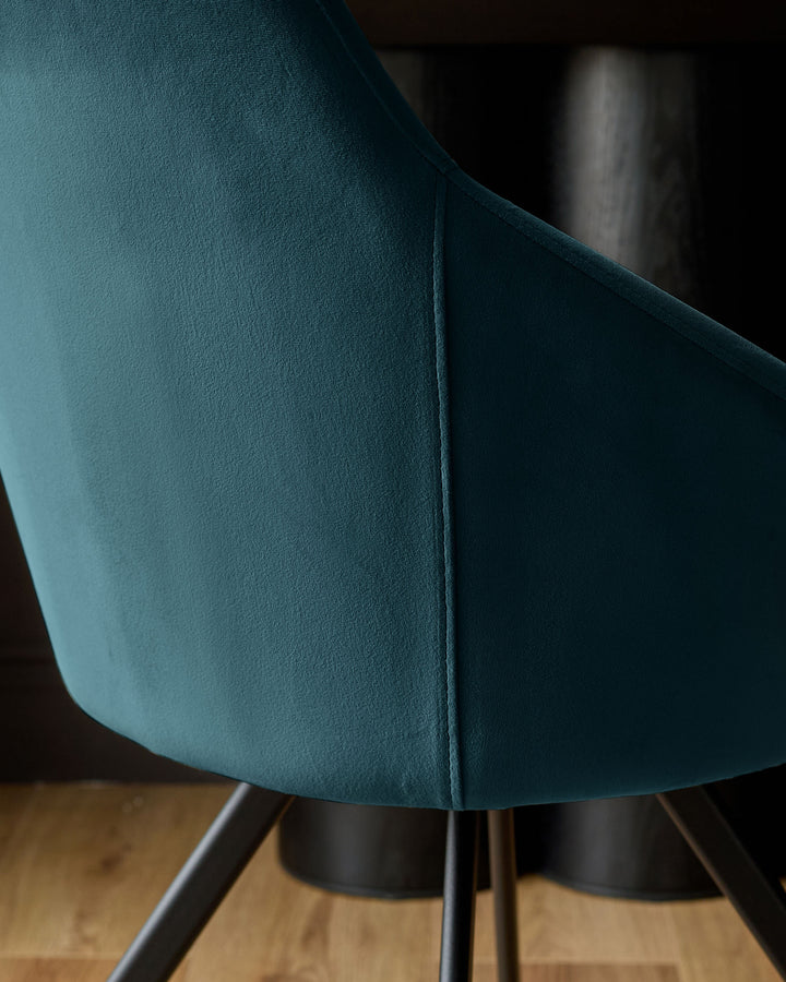 Dark teal velvet swivel chair, close-up detail showing curved backrest seam and black metal base legs on wooden floor.
