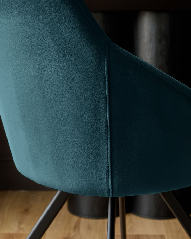 Dark teal velvet swivel chair, close-up detail showing curved backrest seam and black metal base legs on wooden floor.
