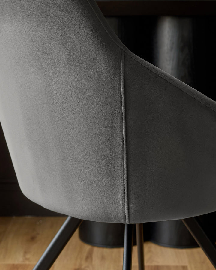 Cleo Velvet Swivel Chair in dark grey, close-up detail showing curved backrest seam and black metal base legs on wooden floor.