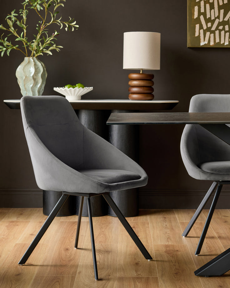 Cleo dark grey velvet swivel chair with curved bucket seat and angled black metal legs, shown in dining setting.