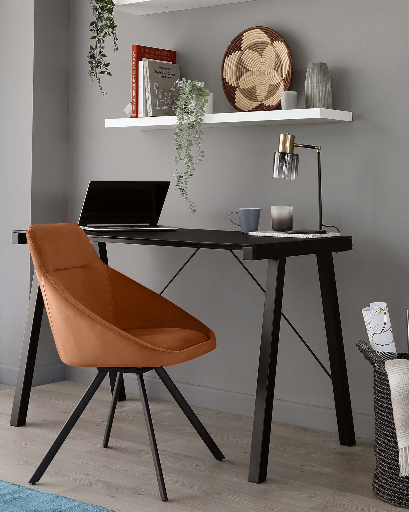 A contemporary home office setup featuring an angular black desk with a laptop on top, paired with a burnt orange upholstered desk chair with black legs. A wall-mounted white shelf displaying books and decorative items hangs above the desk, and a modern gold and black desk lamp illuminates the workspace.