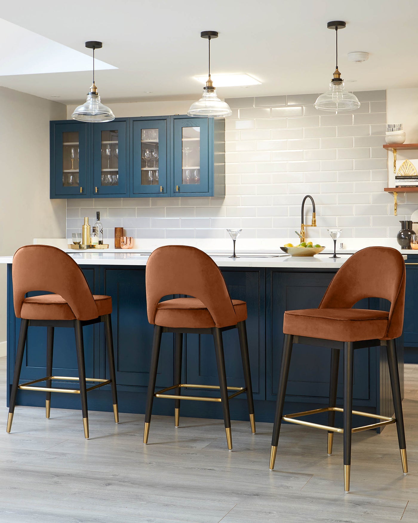 Three modern bar stools with sleek design, featuring burnt orange velvet upholstered seats and backrests, black legs with brass accents, and a footrest.