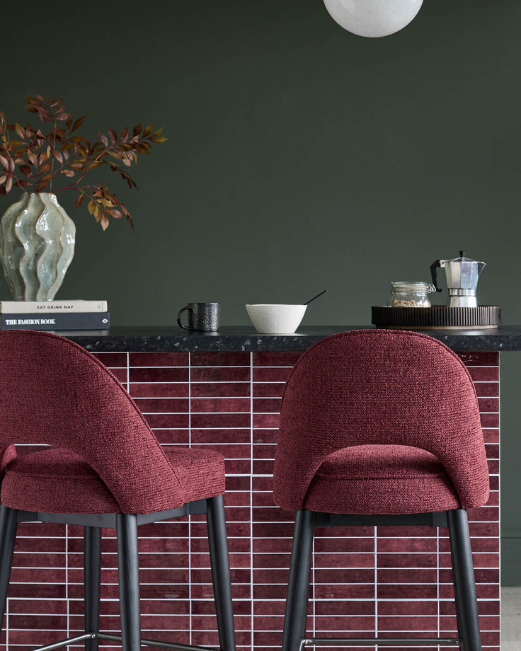 Two bar stools with red fabric seats and black legs sit at a red tiled breakfast bar against a dark green wall.
