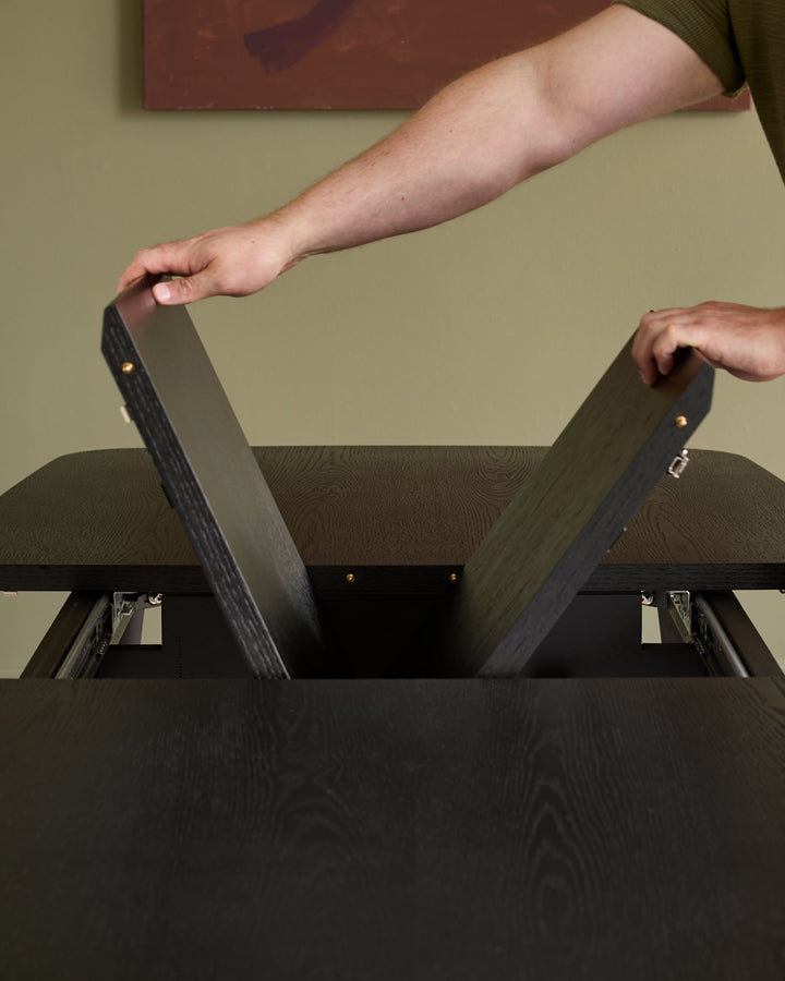 Black wood extending dining table with leaf extension being pulled open by two hands, showing metal hardware and extension mechanism.