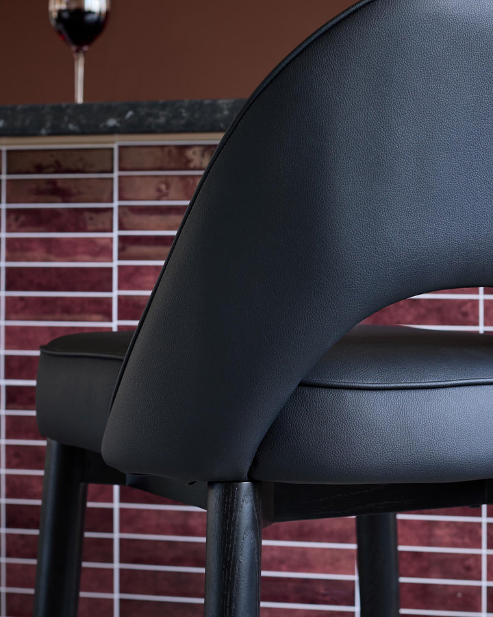 A close-up of a modern black bar stool with dark wooden legs, set against a brick-effect tiled wall, suggesting stylish home decor.