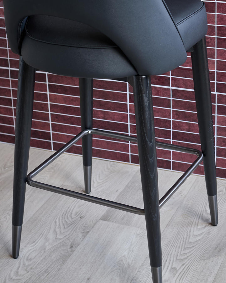 Close up of a modern black bar stool with dark wood legs and a metallic footrest on a light wooden floor.
