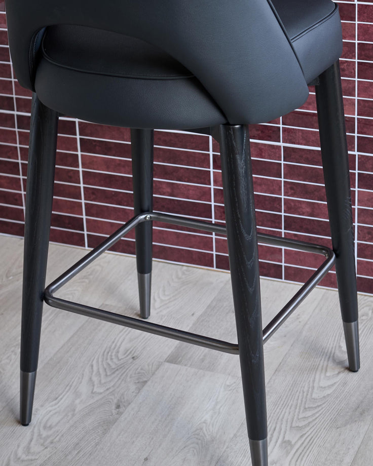 Close up of a modern black bar stool with dark wood legs and a metallic footrest on a light wooden floor.