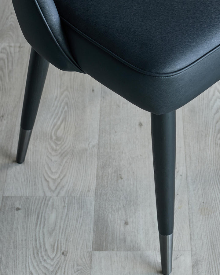 Close-up of a sleek, modern chair with black faux leather seat and dark grey legs with silver caps, on a light wood-effect floor.