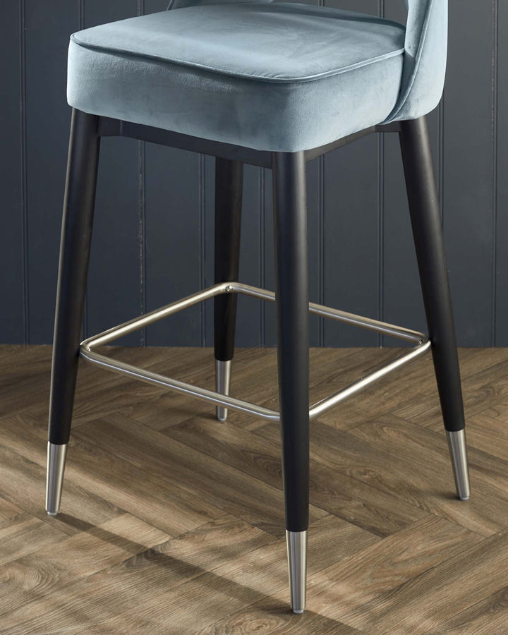 Elegant modern bar stool with a plush velvet cushion seat in a muted blue tone, featuring a supportive curved backrest. The stool has sleek black tapered legs with metallic silver tips and a matching footrest, combining comfort with a contemporary chic design.