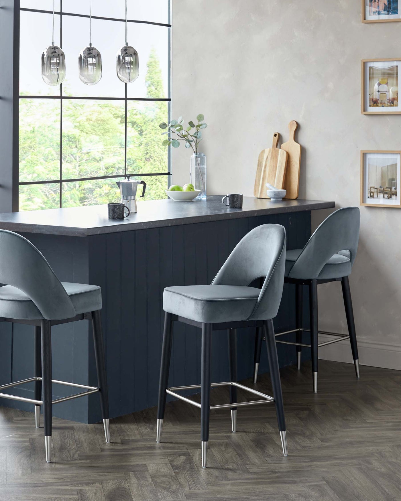Modern grey velvet bar chairs with black legs and metallic footrests in front of a kitchen island with a black base and a grey countertop in a stylish kitchen setting.