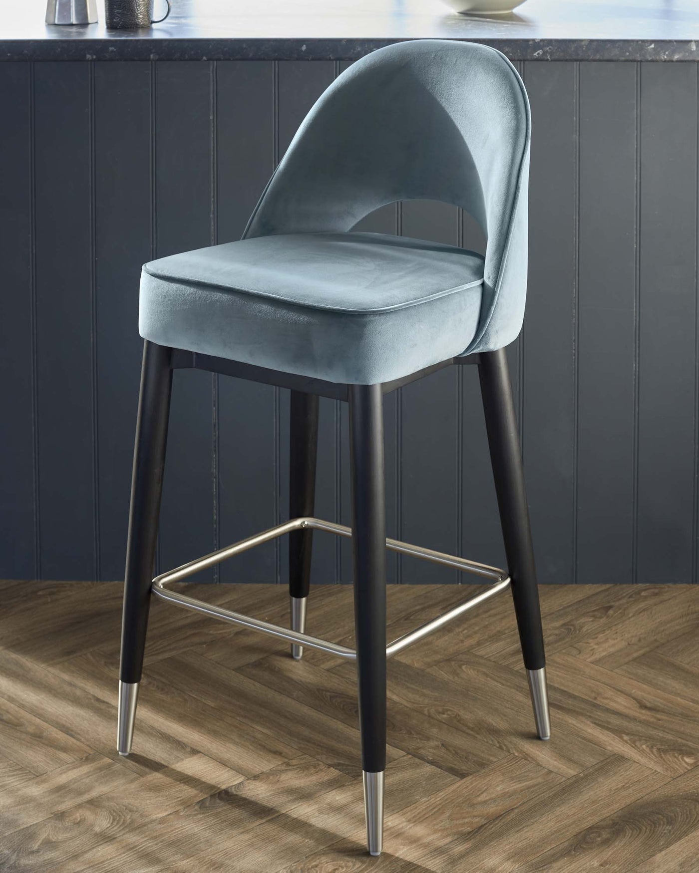 Elegant contemporary bar stool with plush teal velvet upholstery, featuring a curved backrest and a padded seat for comfort. The stool stands on tapered black legs accented with sleek metallic tips and a matching footrest, creating a stylish contrast. Perfect for modern kitchen or bar settings.