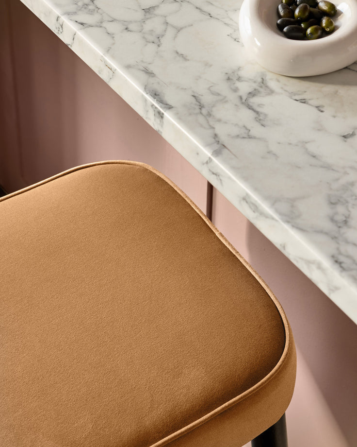 Caramel fabric backless bar stool with curved seat and piping detail, shown beside marble countertop.
