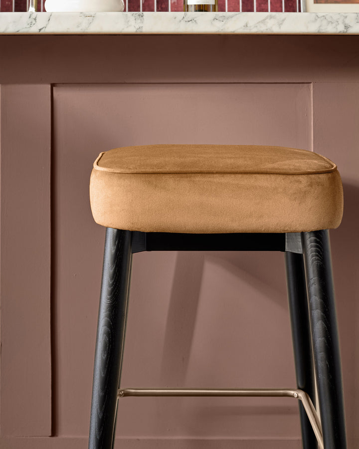 Clover backless bar stool with caramel fabric seat and black wooden legs with brass footrest.