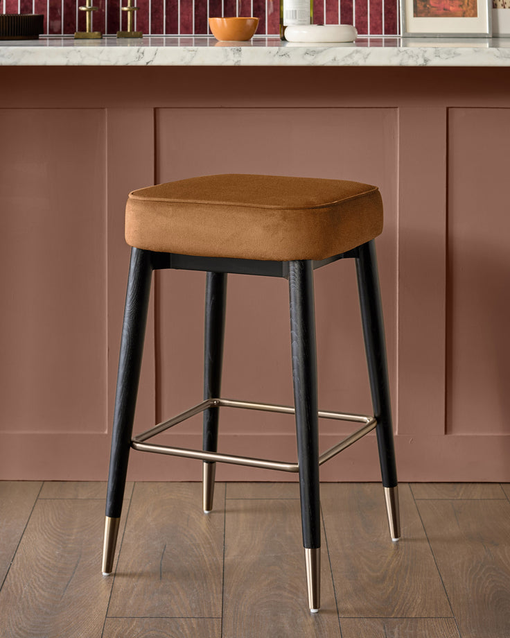 Clover backless bar stool in caramel fabric with black wooden legs and brass foot caps, shown against mauve paneled wall.