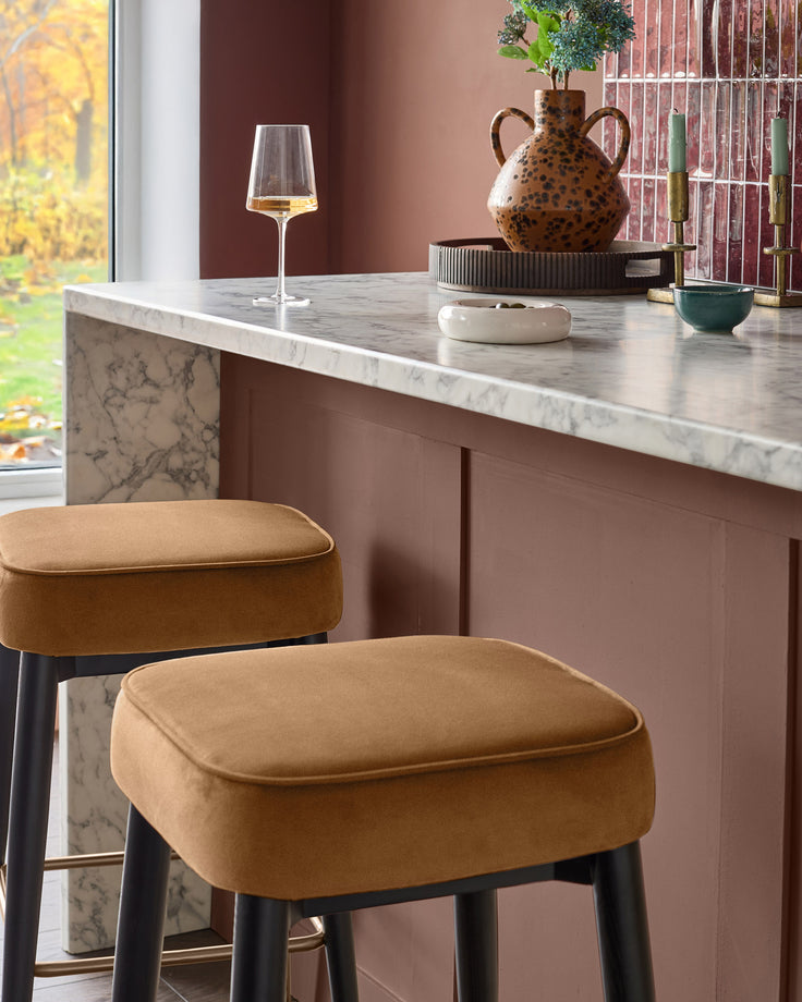 Caramel fabric backless bar stool with black legs, shown at marble kitchen counter in modern room setting.