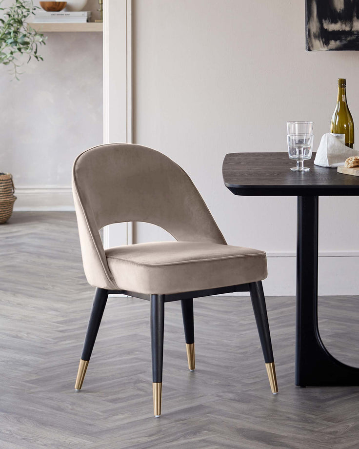 A modern chair in taupe velvet with black and gold legs sits next to a dark wood dining table.