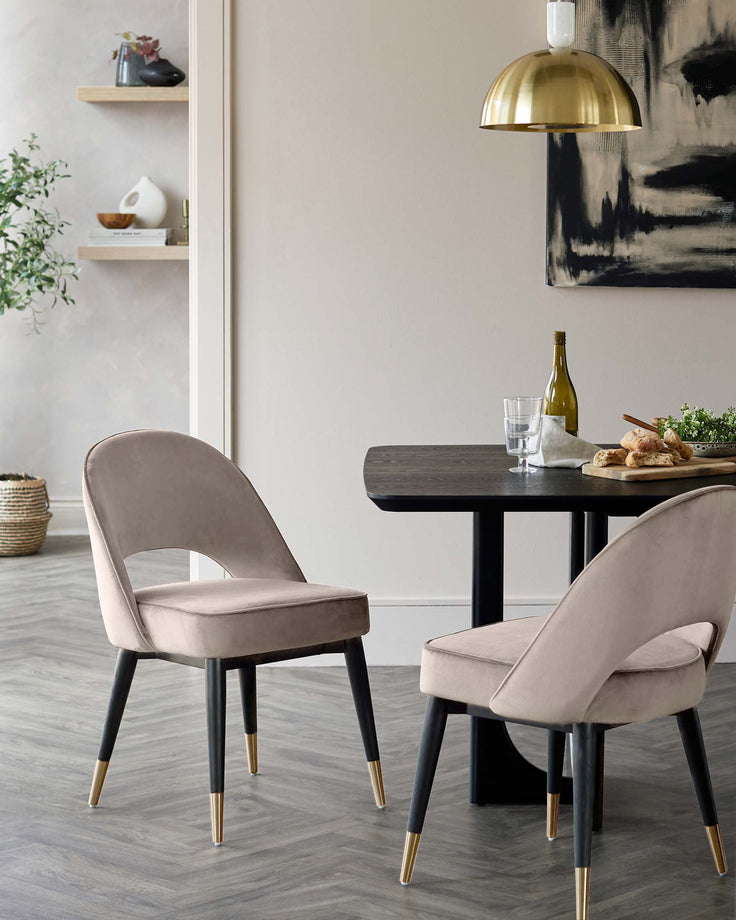 Two velvet chairs with black legs and gold tips sit at a dark wooden table, illuminated by a gold pendant light.