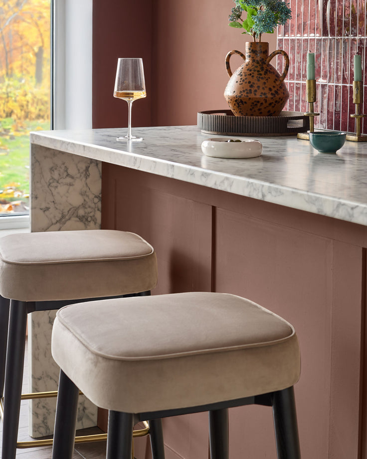 Clover Velvet Backless Bar Stool in Champagne with black legs, shown at marble counter in modern interior setting.