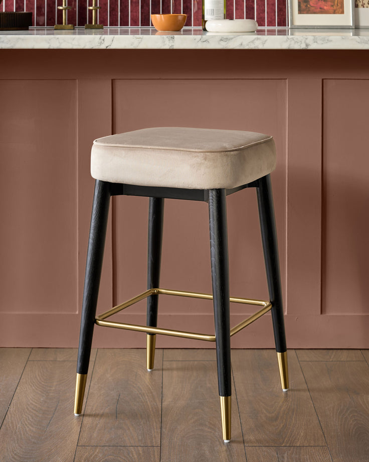 Clover Velvet Backless Bar Stool in Champagne with black wooden legs and brass footrest, shown against mauve paneled wall.