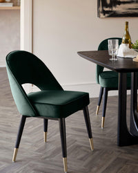Close-up of two stylish, green velvet dining chairs with black legs and gold feet, positioned around a dark wood table.