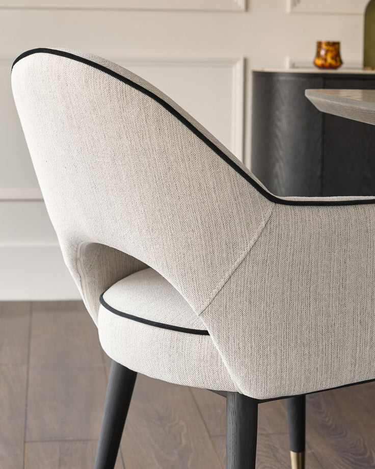Clover dining armchair in ivory fabric, side angle view showing curved backrest with black piping detail and dark wooden legs.