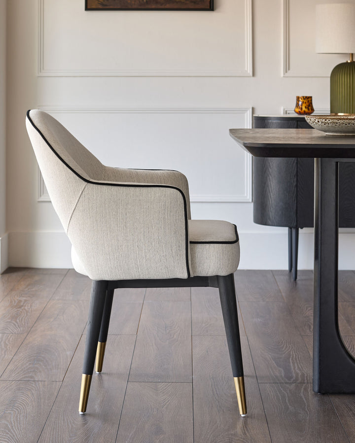 Clover dining armchair in ivory fabric, angled back view showing black piping trim and brass leg caps against wood flooring.