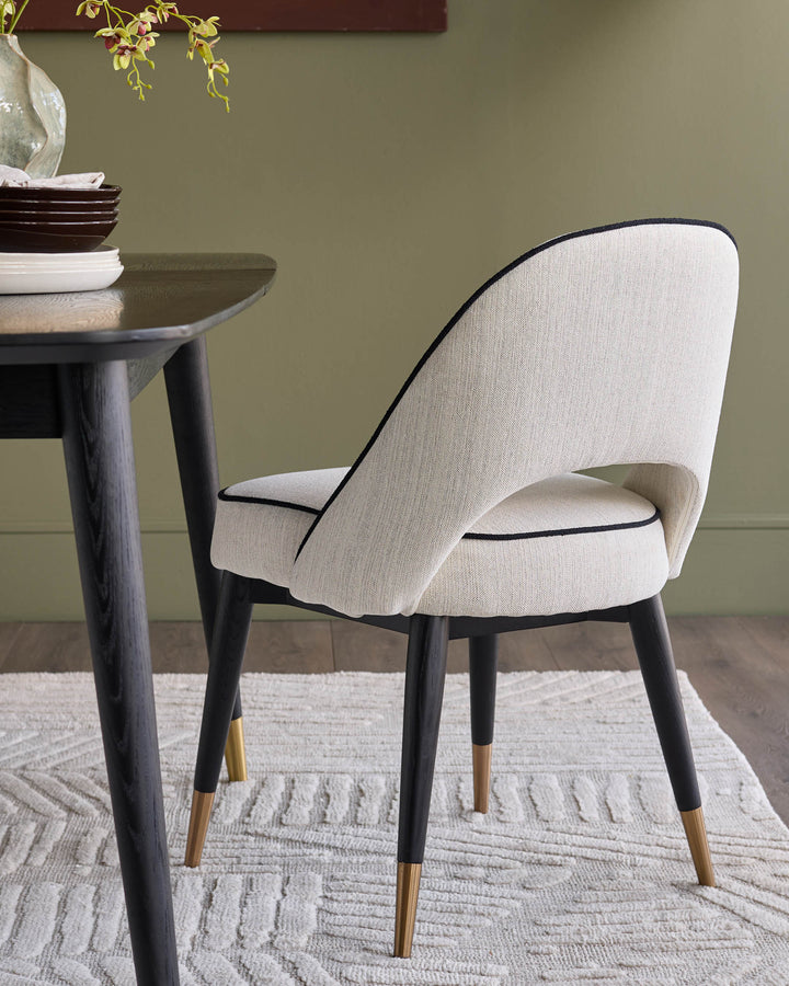 Clover ivory fabric dining chair with black piping, dark wood legs, and brass foot caps, shown at a table setting.