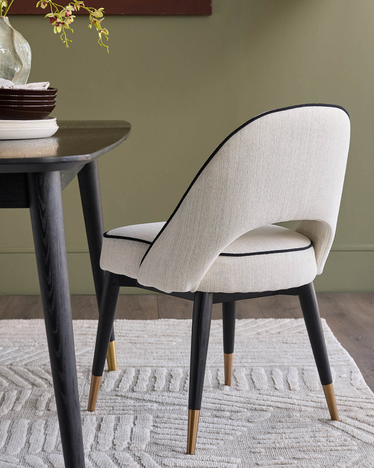 Clover ivory fabric dining chair with black piping, dark wood legs, and brass foot caps, shown at a table setting.