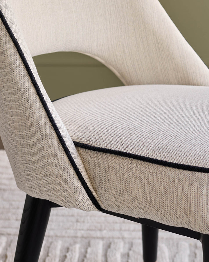 Clover fabric dining chair in ivory, angled side view showing curved backrest, black piping detail, and dark wooden legs.