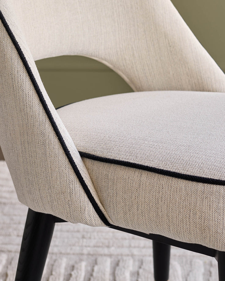 Clover fabric dining chair in ivory, angled side view showing curved backrest, black piping detail, and dark wooden legs.