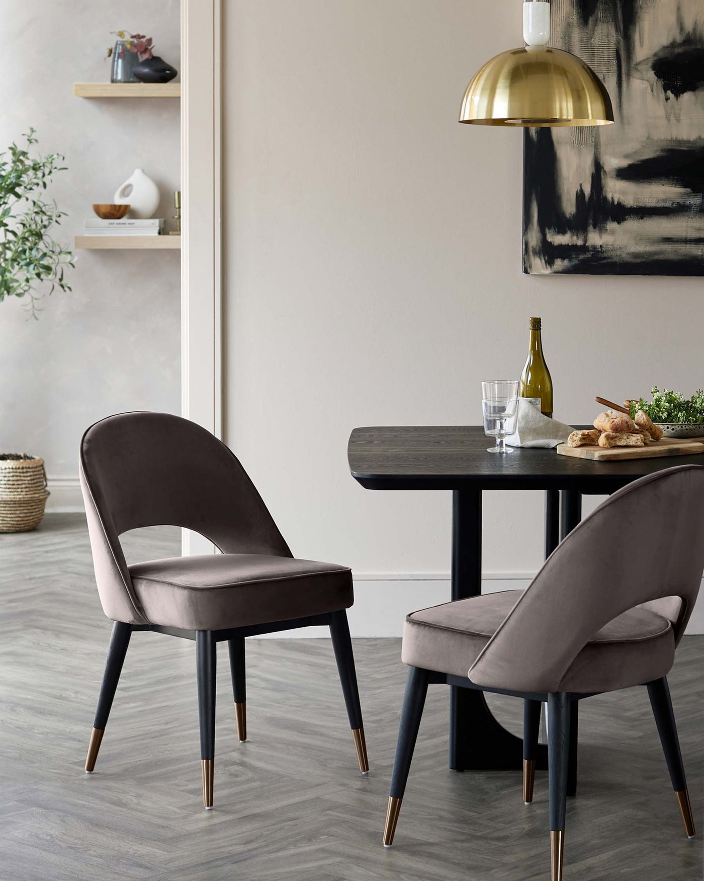 A modern dining set featuring two plush, mauve upholstered chairs with black legs accented with gold tips, surrounding a dark-stained wooden table. The setting is complemented by a large brass-domed pendant light overhead and minimalistic shelf decor against a neutral wall.