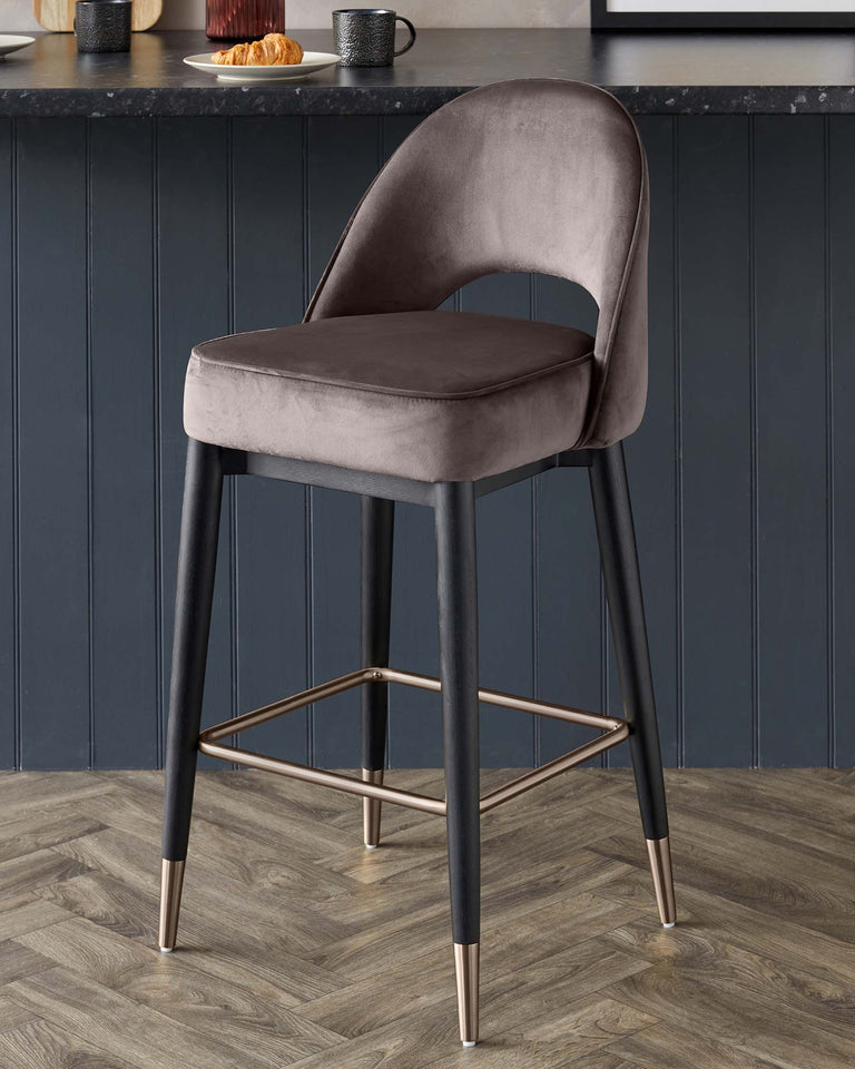 Elegant modern bar stool with a curved, chair-like backrest and plush seat upholstered in a soft grey velvet fabric. The stool features an angular metal frame with a matte black finish and contrasting gold tips on the legs, providing a chic and contemporary aesthetic. Perfect for adding a touch of sophistication to any kitchen or dining space.