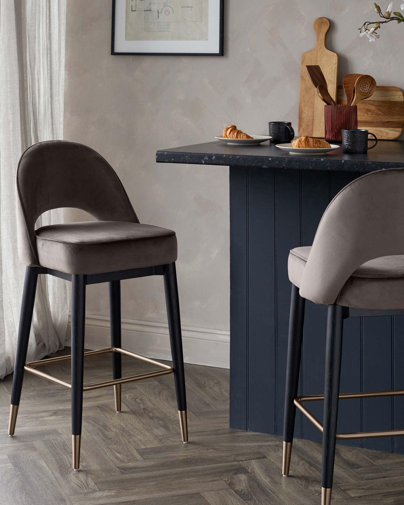 Two elegant, modern bar chairs with dark brown upholstery and black frames featuring gold-coloured accents on the leg tips and footrests. The chairs are positioned at a black kitchen counter that displays a simple breakfast setup.