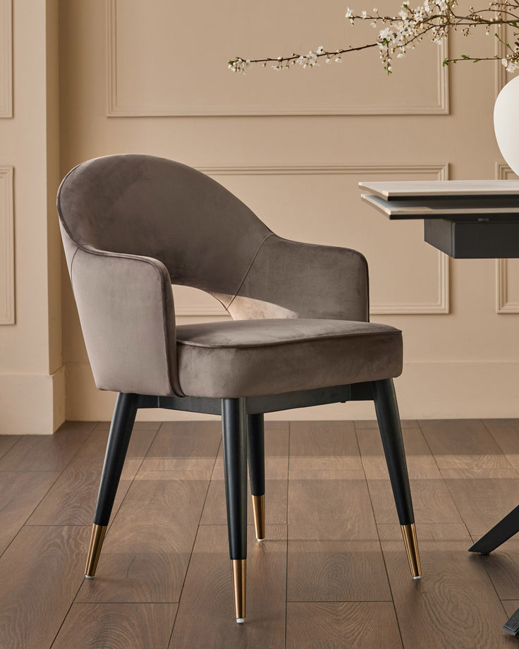 Mocha velvet dining armchair with curved back, black wooden legs, and gold-tone foot caps, shown in a styled interior setting.