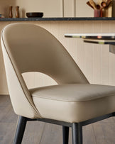 Beige leather dining chair with wooden legs, next to a dining table. Stylish seating for your dining space.