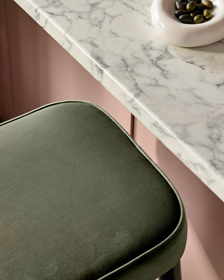 Clover Velvet Backless Bar Stool in Olive Green, shown at an angle against a pink wall and marble countertop with olives.