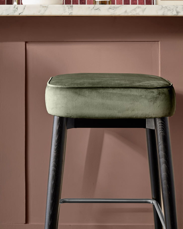 Clover velvet backless bar stool in olive green with dark wooden legs and footrest against mauve wall.