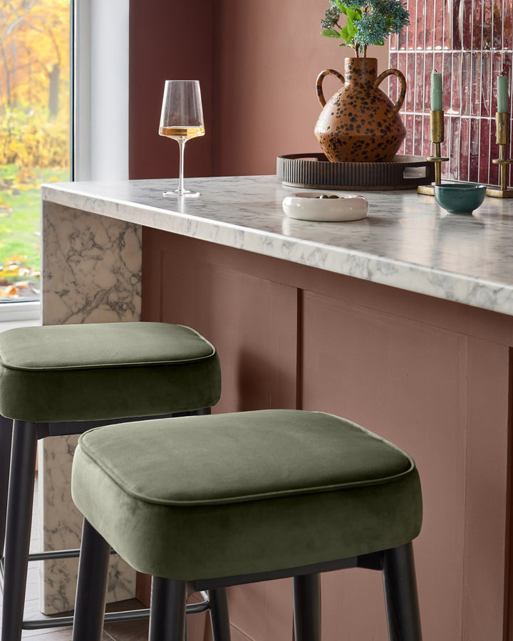 Clover Velvet Backless Bar Stool in Olive Green with black legs, positioned at a marble countertop in a styled kitchen setting.
