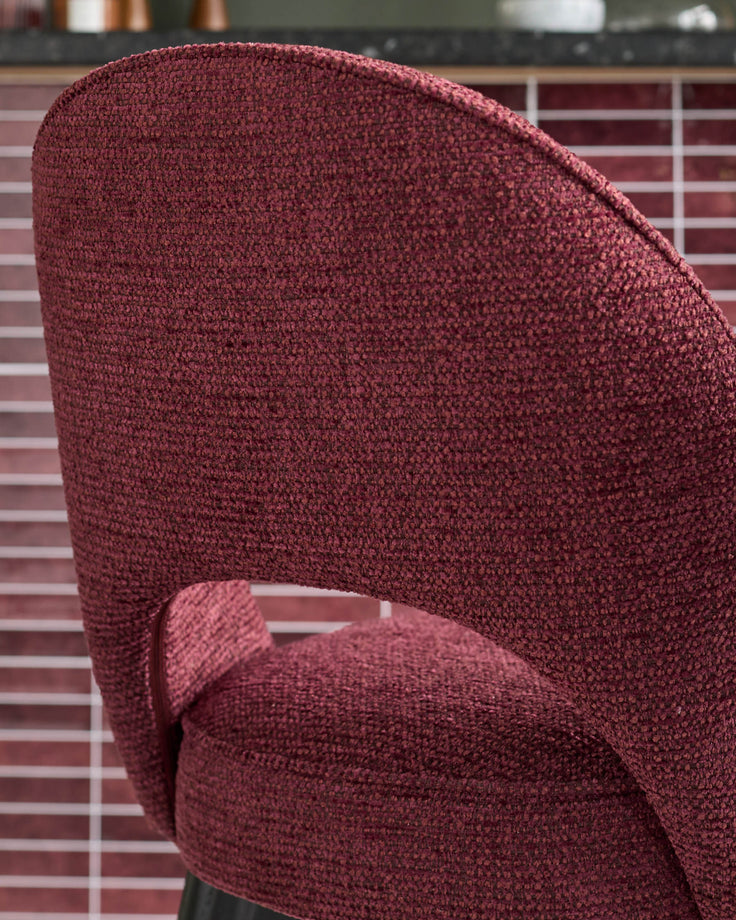 Close-up of a stylish burgundy fabric chair, showing its textured upholstery and curved back design against a tiled wall.