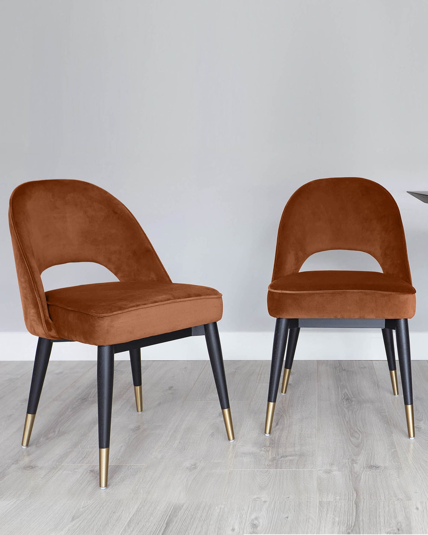 Two contemporary velvet dining chairs with a rich caramel hue, featuring curved backrests and cushioned seats, supported by sleek, tapered legs in a black finish with elegant gold-tone metal tips.