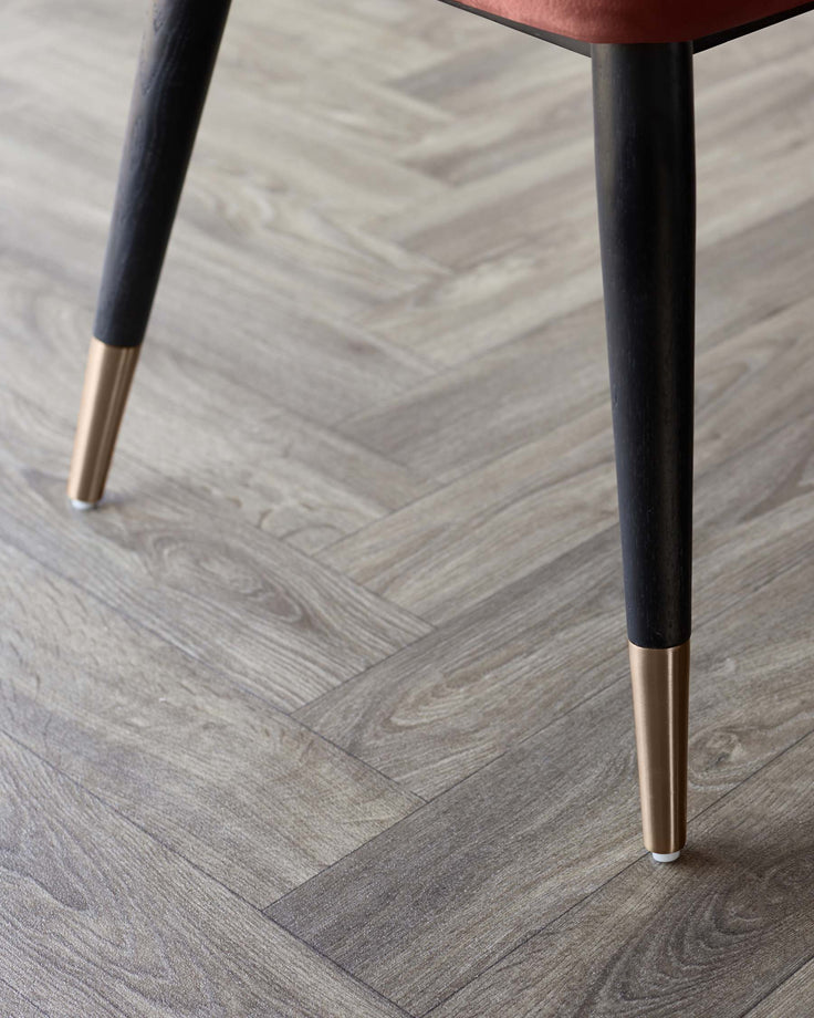 Close up of a chair leg with brass foot, on a herringbone grey wood floor. Shows attention to detail and luxury materials.