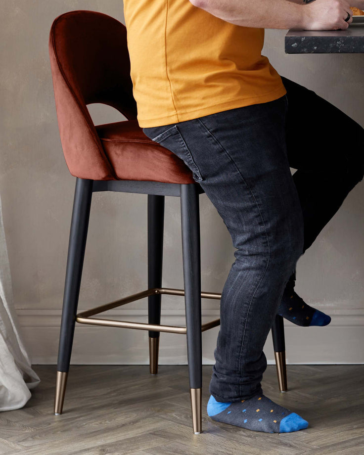A modern bar stool with a dark wood frame, copper accents, and a luxurious velvet seat in an autumnal shade of orange.