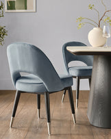 Clover velvet dining chair in blue with curved backrest and black legs with stainless steel caps, shown at table.