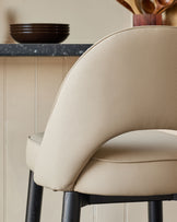 Close up of a bar stool with a cream leather seat and back and black legs.