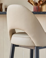 A cream leather bar stool with a rounded back and dark wood legs sits in a kitchen. The focus is on the stool's design and textures.