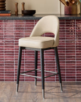 A modern bar stool with a cream seat and black legs, featuring a silver footrest, stands against a red tiled backdrop.