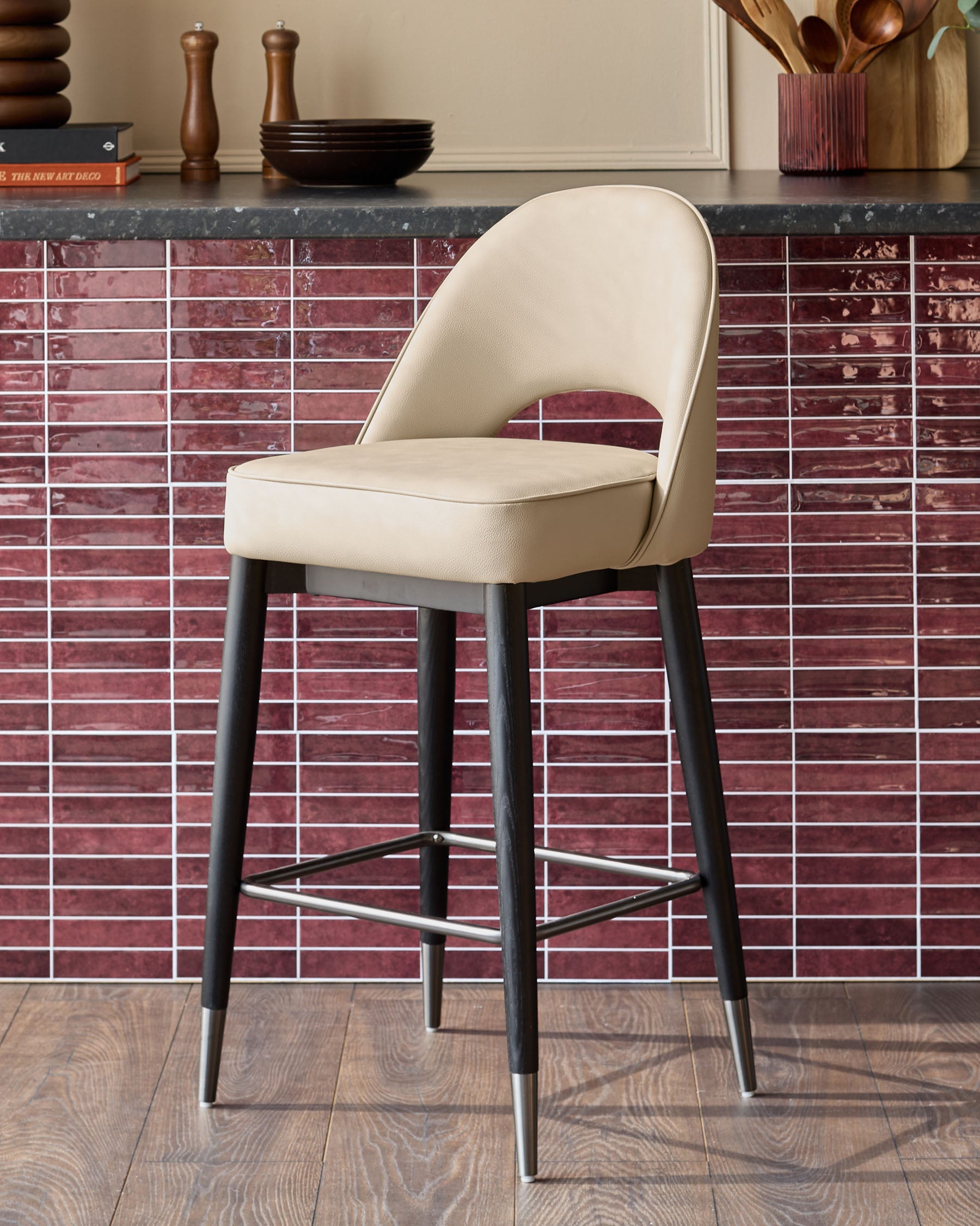 A modern bar stool with a cream seat and black legs, featuring a silver footrest, stands against a red tiled backdrop.